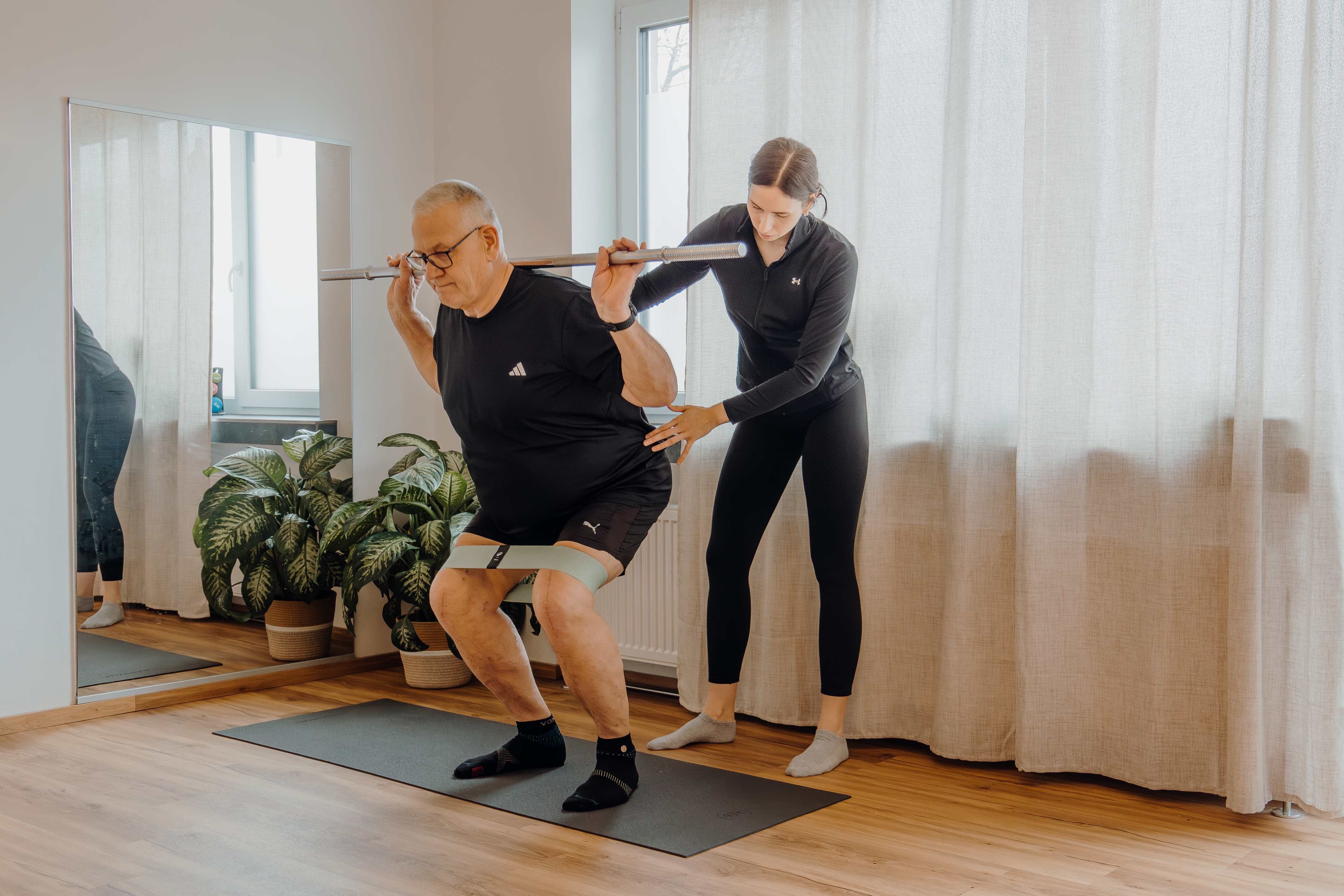 Patient bei Kniebeugen mit Langhantel, angeleitet von Therapeutin